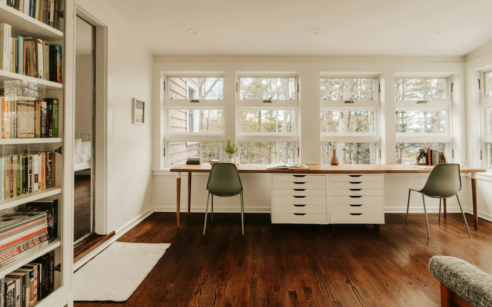 A bright study room with a wall of bookshelves, a long wooden desk and two chairs facing tall windows onto trees.