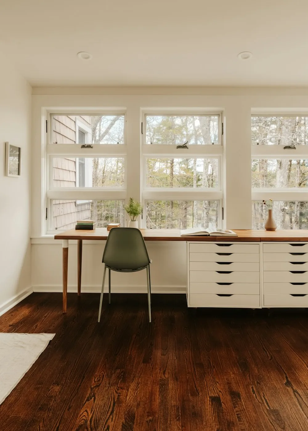 A bright study room with a wall of bookshelves, a long wooden desk and two chairs facing tall windows onto trees.